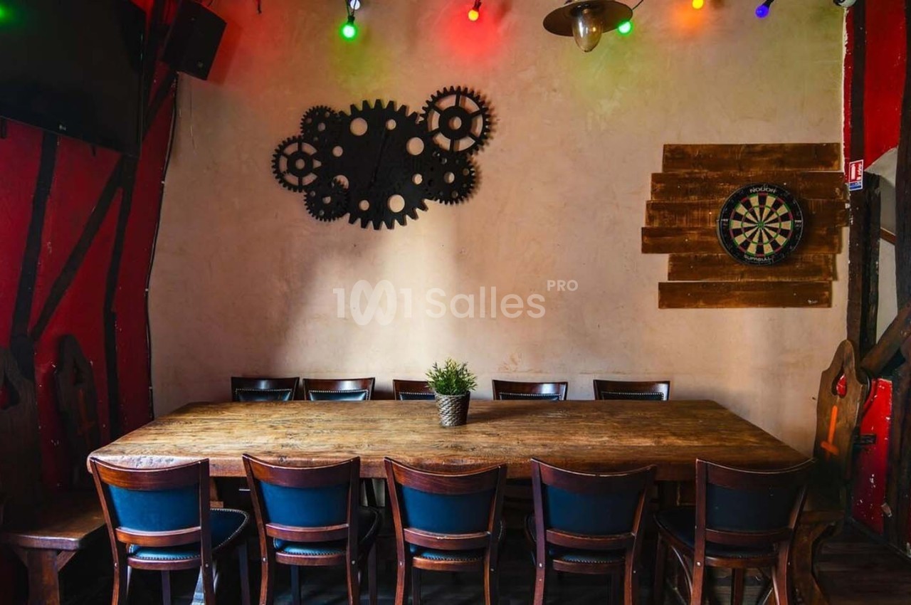 Salle avec une grande table en bois entourée de chaises, décorée de guirlandes lumineuses et d'engrenages au mur.