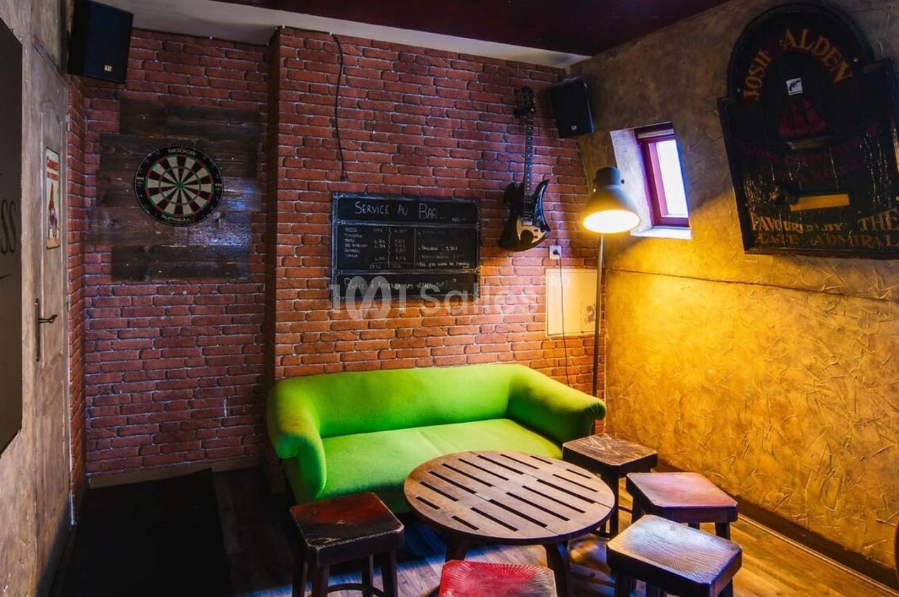 Salle conviviale avec murs en briques, canapé vert, table basse, tabourets, cible de fléchettes et éclairage tamisé.