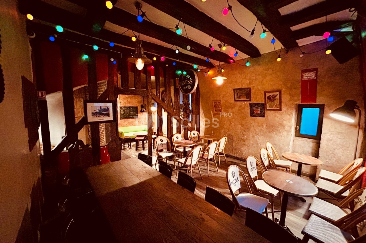 Salle chaleureuse avec tables en bois, chaises disposées, murs en pierre et guirlandes lumineuses colorées suspendues.