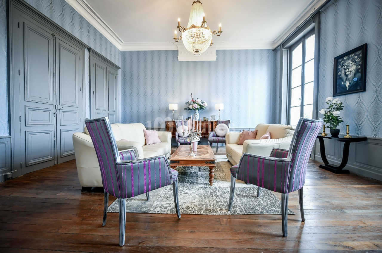 Salon élégant avec mobilier classique, fauteuils colorés, table basse en bois, lustre et lumière naturelle.