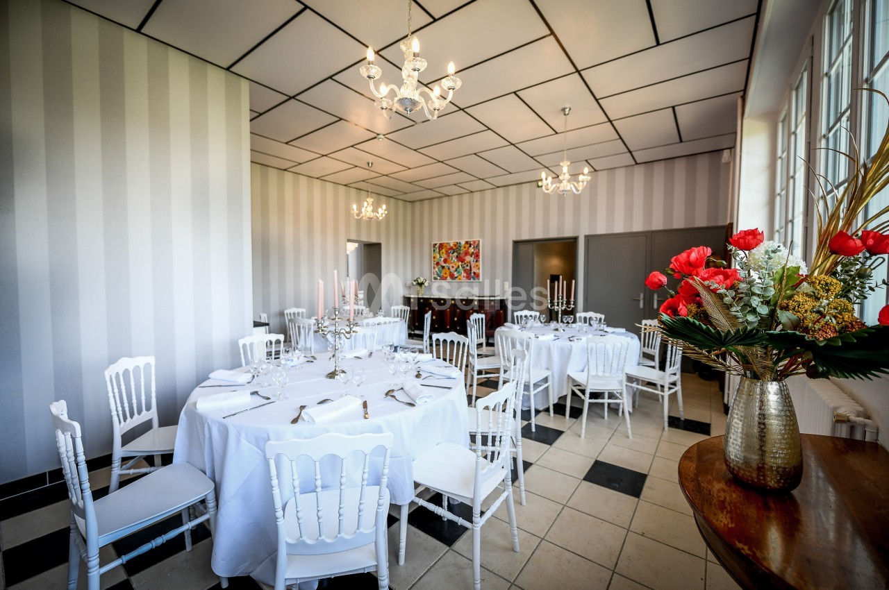 Salle lumineuse avec tables rondes dressées, chaises blanches, lustres et décoration florale dans un cadre élégant.