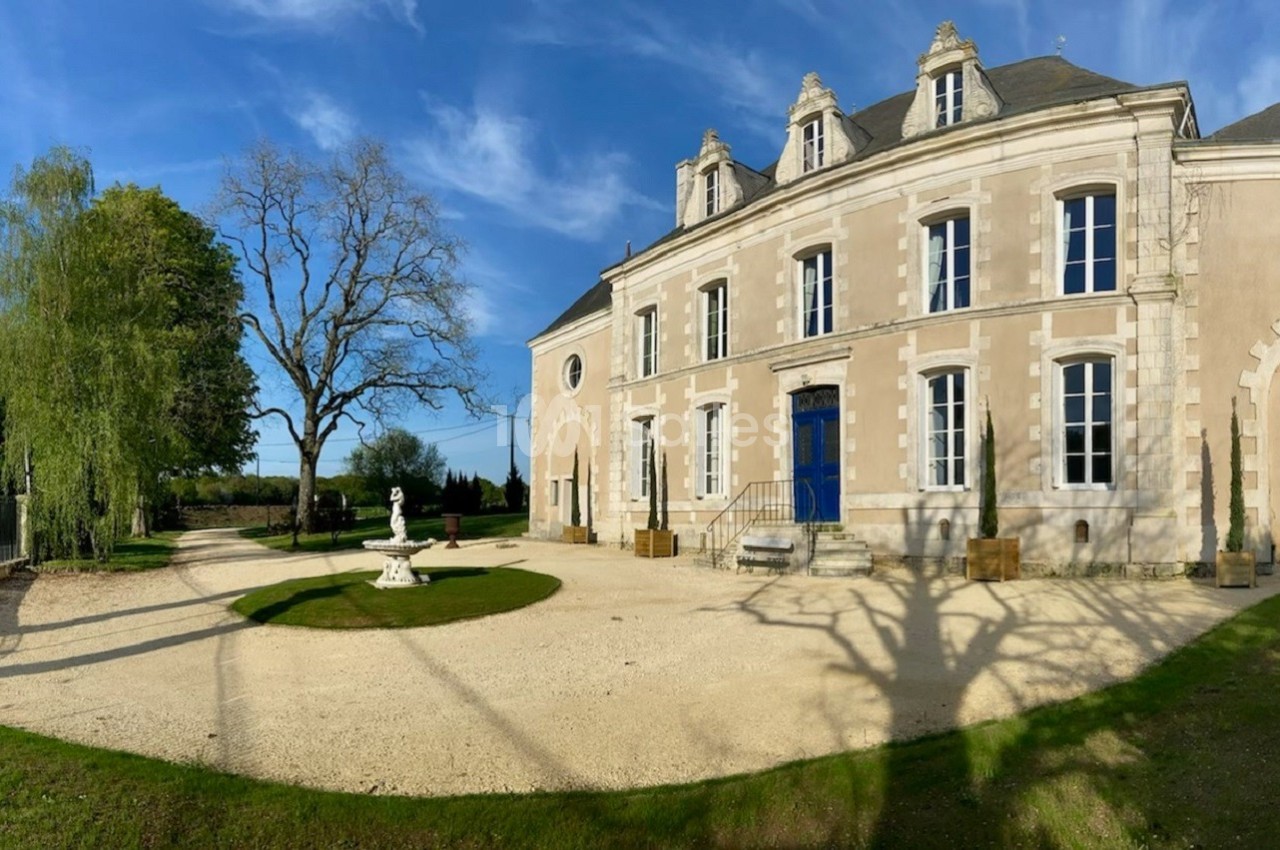 Façade d'un manoir en pierre avec une porte bleue, entouré d'un jardin arboré et d'une allée gravillonnée.