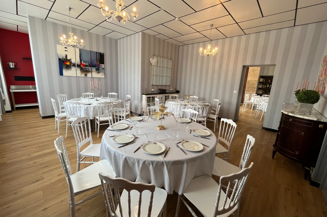 Salle de réception élégante avec tables rondes dressées, chaises blanches et lustres suspendus.