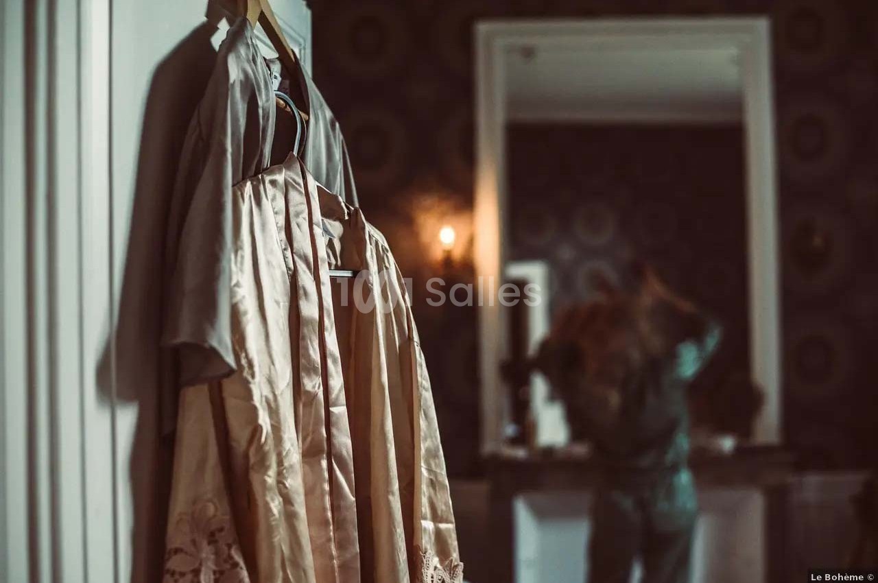 Robe beige suspendue à un cintre au premier plan, avec une personne floue se coiffant devant un miroir en arrière-plan.