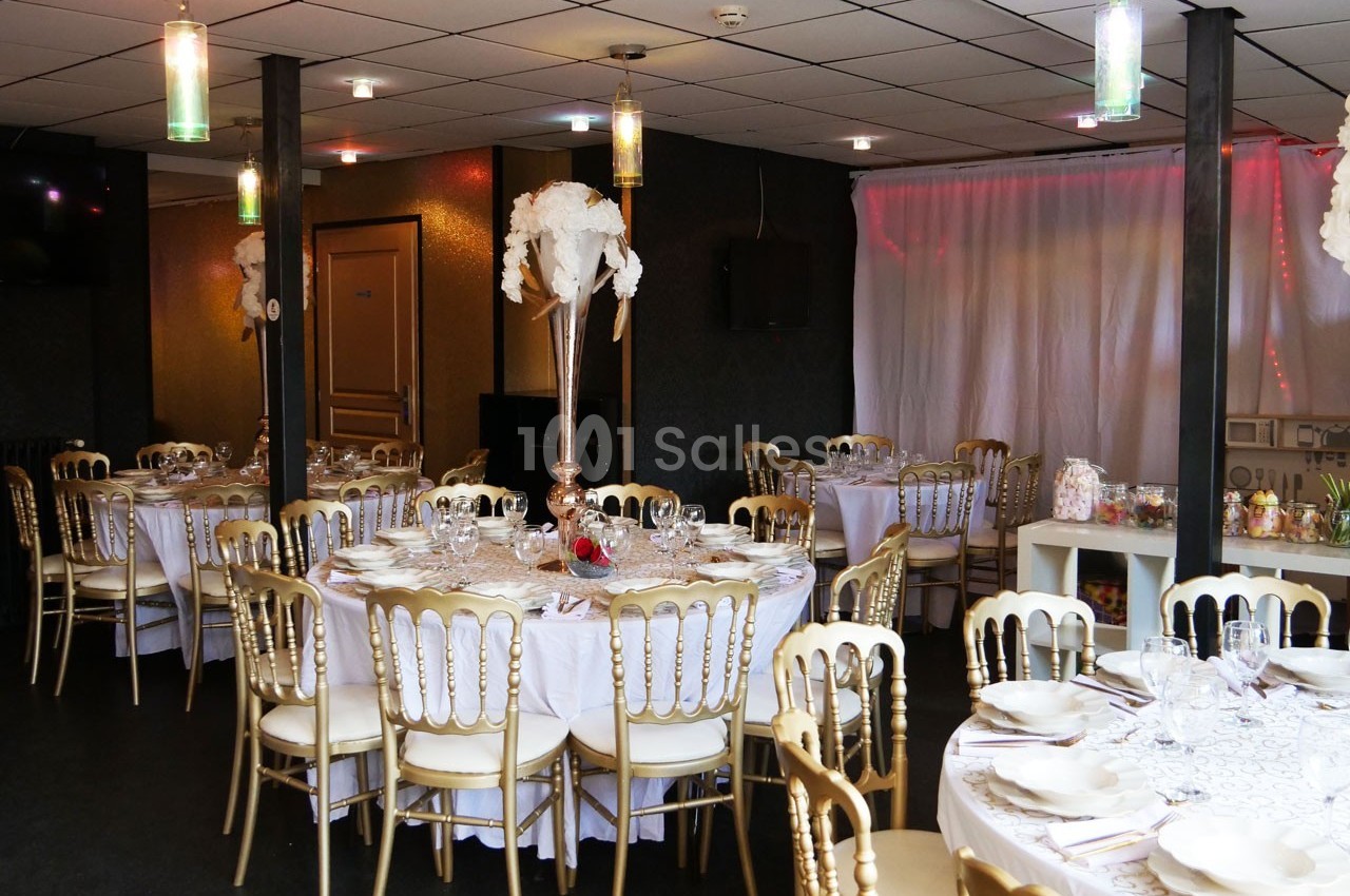 Salle de réception décorée avec des tables rondes dressées, chaises dorées et centre de table floral blanc.