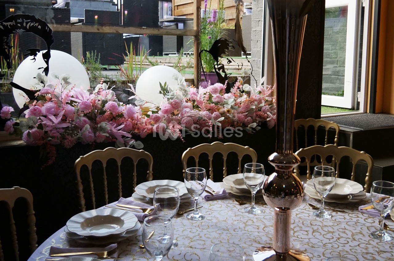 Table dressée avec vaisselle élégante, centre de table argenté et décor floral rose près d'une baie vitrée.