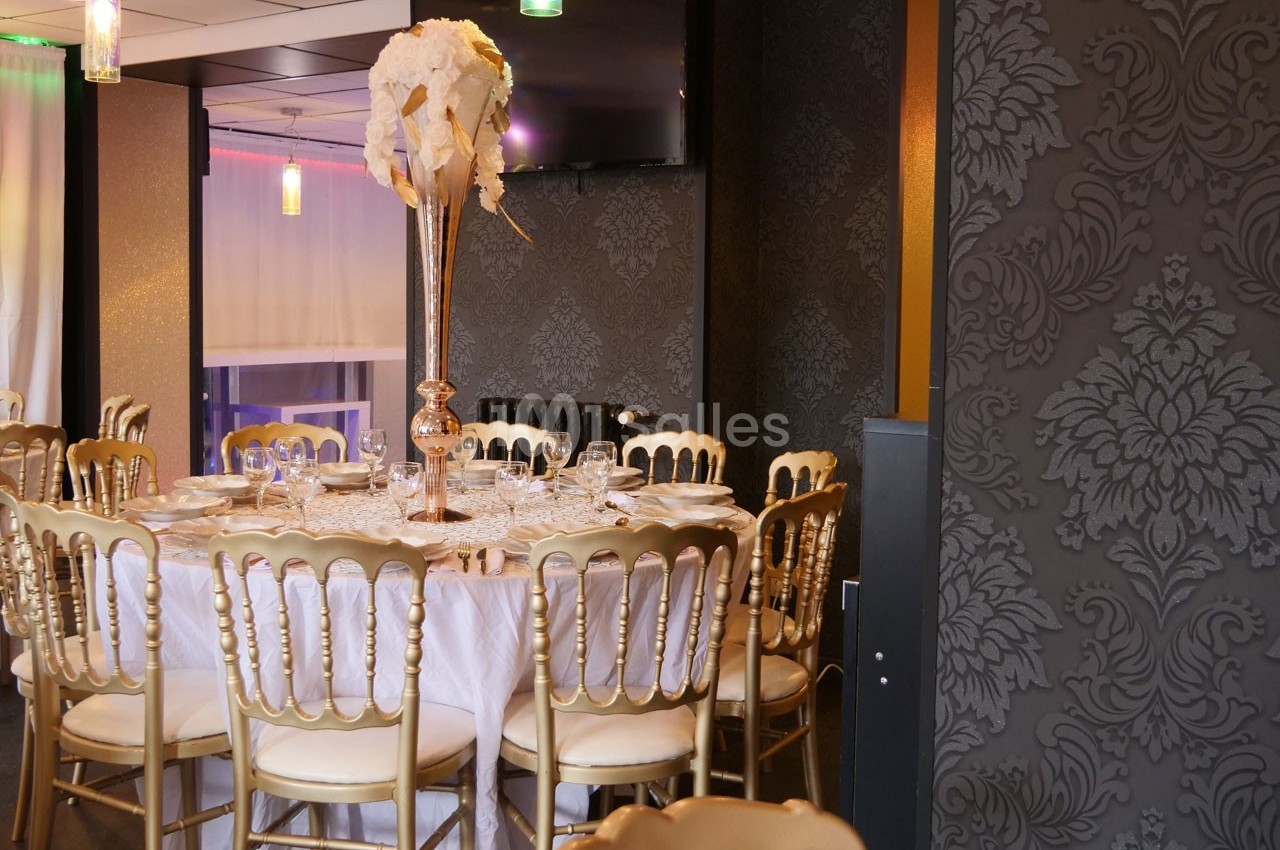 Salle de réception élégante avec table ronde dressée, chaises dorées et décoration florale sur un vase haut.