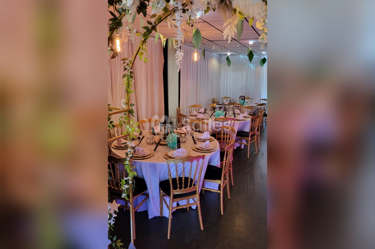 Salle décorée pour un événement avec tables rondes dressées, chaises en bois et guirlandes lumineuses suspendues.