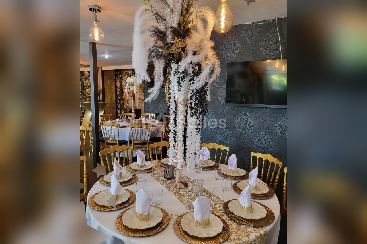 Table élégamment dressée avec nappes blanches, serviettes pliées, vaisselle dorée et centre de table orné de plumes et…