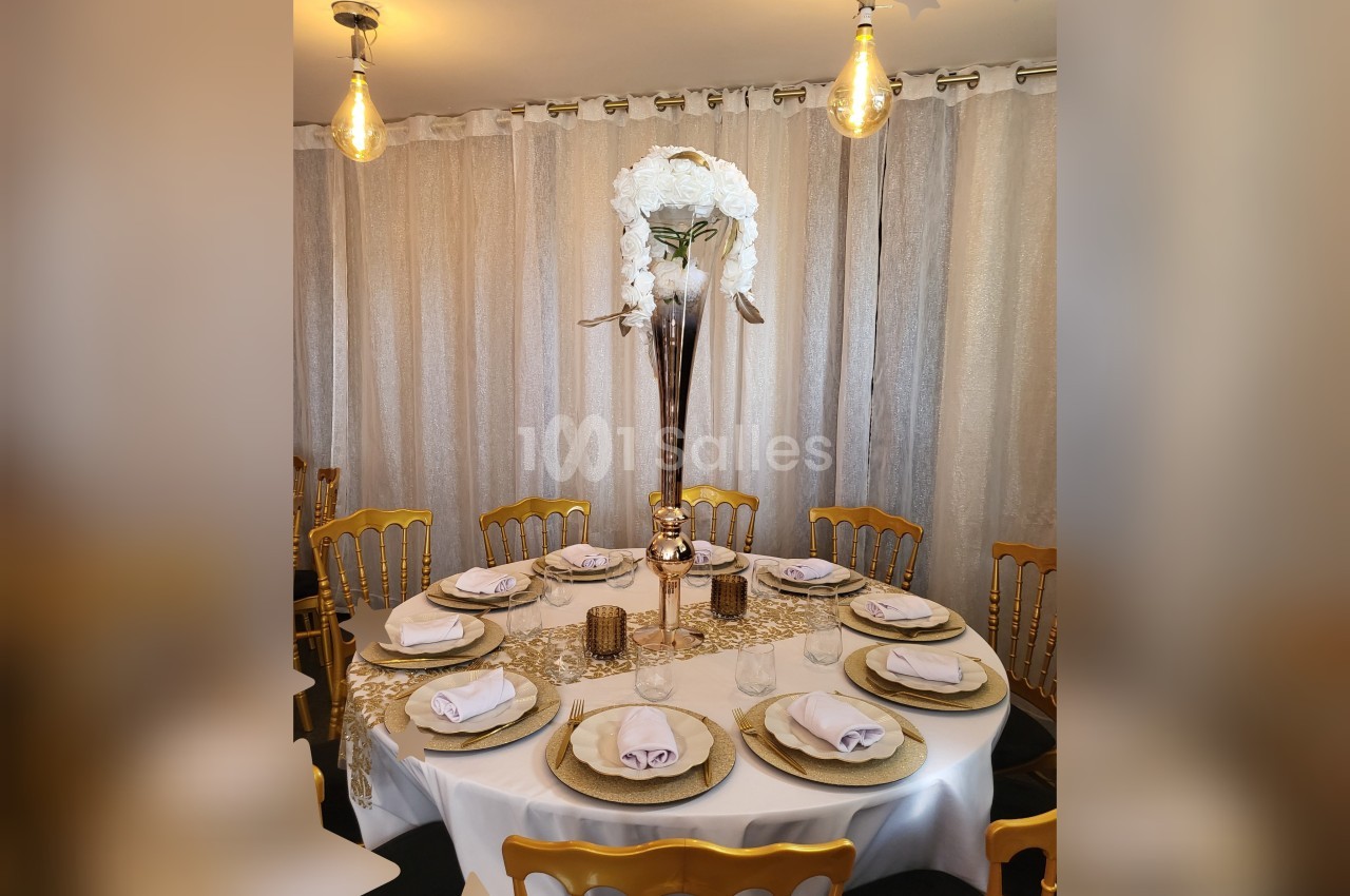 Table ronde élégamment dressée avec des assiettes dorées, des serviettes pliées et un centre de table floral blanc.