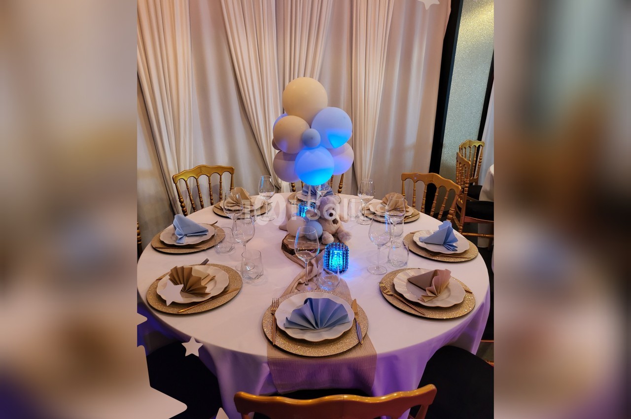 Table décorée pour un événement avec nappes blanches, serviettes pliées, ballons et un ours en peluche au centre.