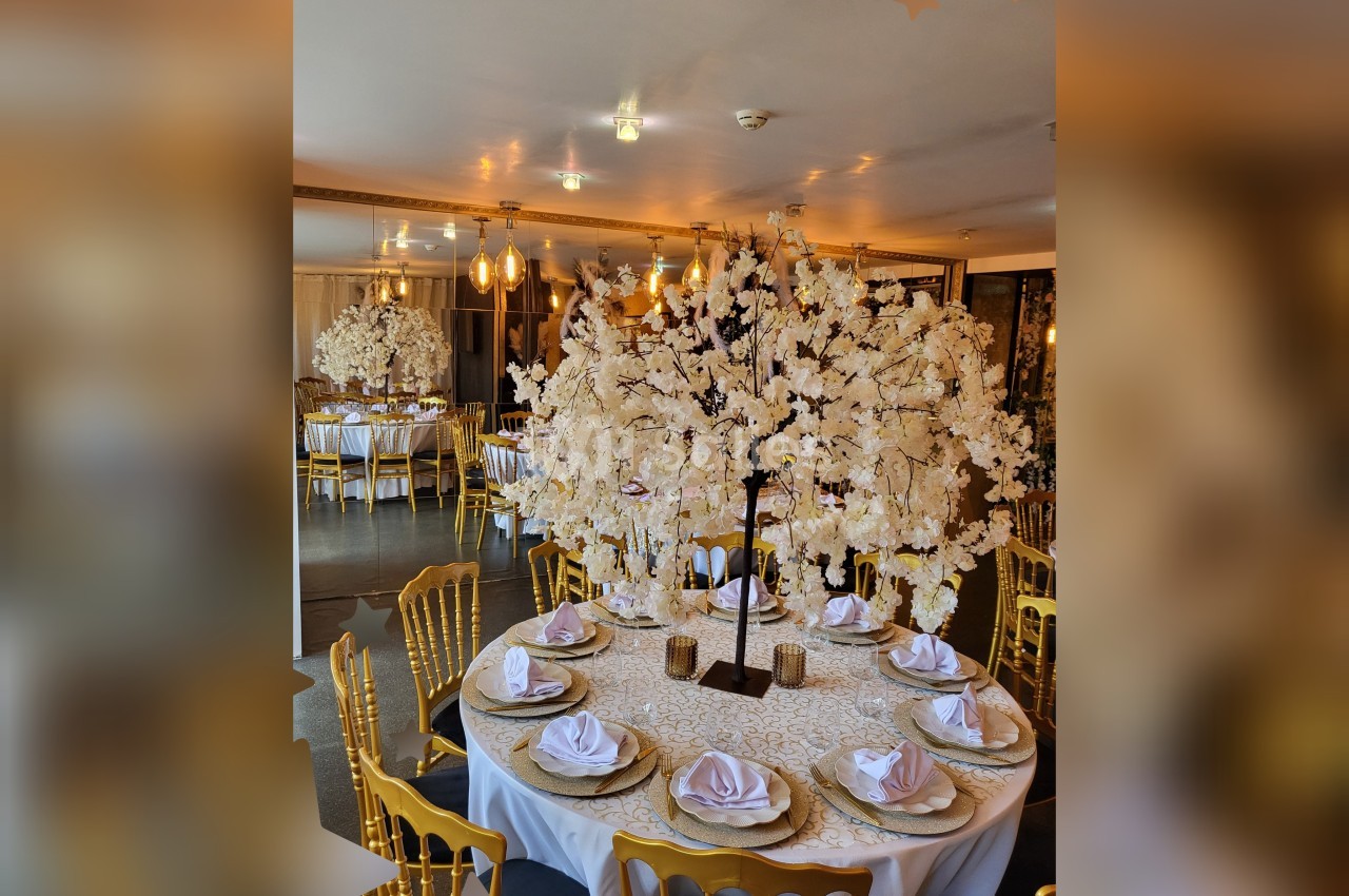 Salle de réception décorée avec des tables rondes, nappes blanches, chaises dorées et centres de table fleuris.