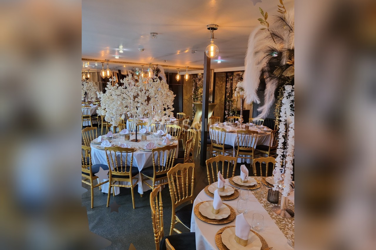 Salle de réception élégamment décorée avec des tables rondes, nappes blanches, chaises dorées et ornements floraux blancs.