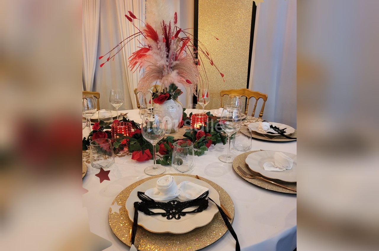 Table élégamment dressée avec assiettes dorées, masques noirs, fleurs rouges et centre de table avec plumes blanches et…