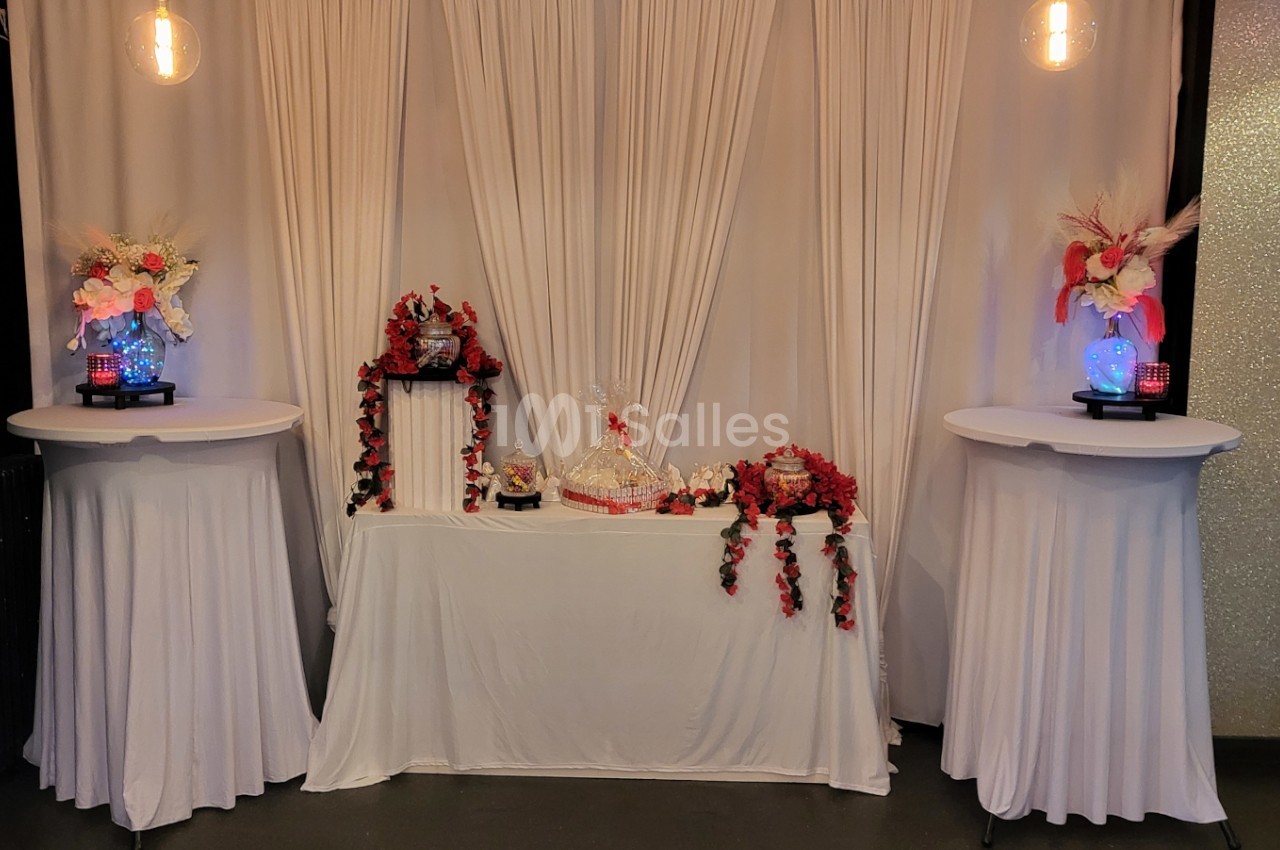 Décoration de table avec nappes blanches, fleurs rouges et roses, et éléments décoratifs sous un fond de rideaux blancs.