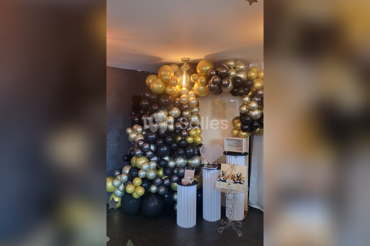 Décoration festive avec arche de ballons noirs, dorés et argentés, entourant des présentoirs blancs dans une pièce sombre.