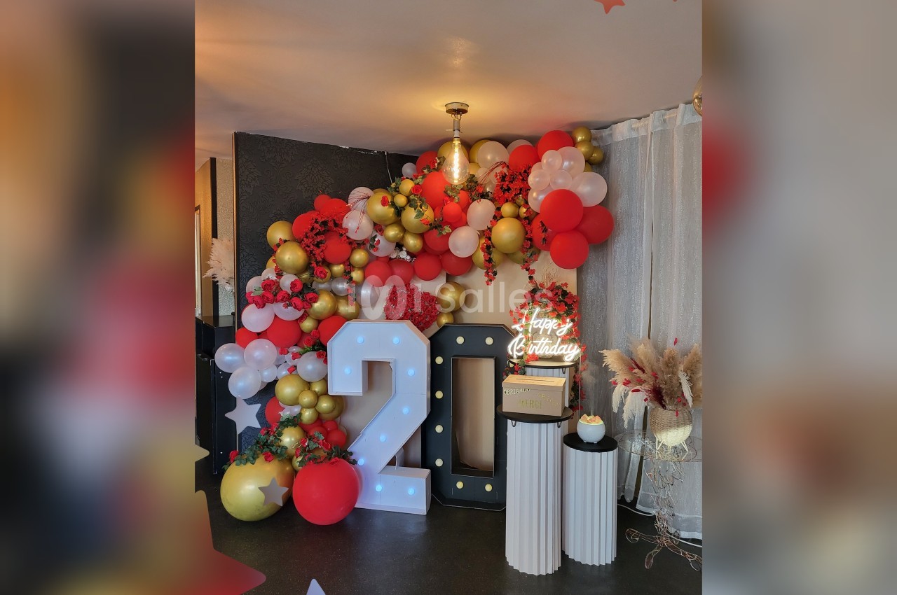 Décoration festive avec ballons rouges, blancs et dorés, chiffres lumineux ’20’ et petit gâteau sur des colonnes.