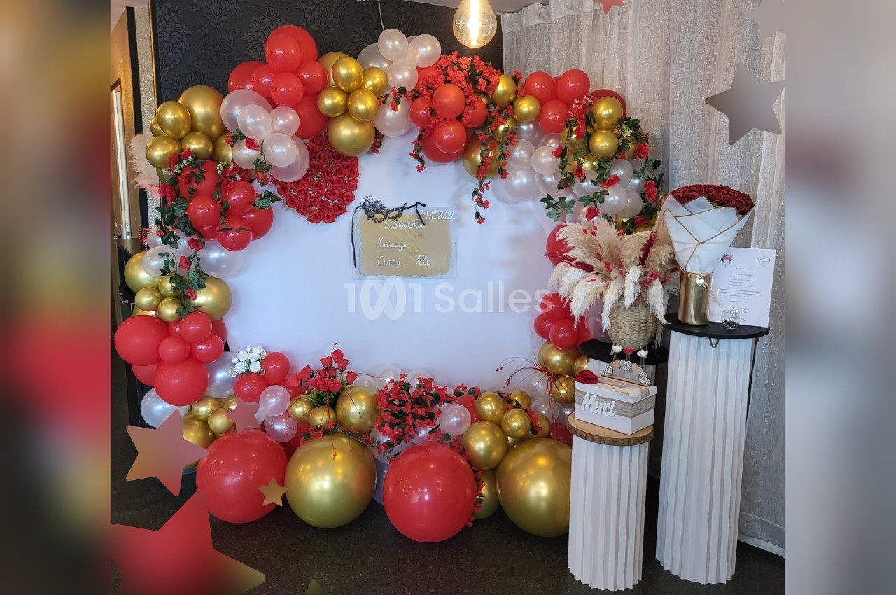 Décor festif avec arche de ballons rouges, dorés et blancs, fleurs, et table avec arrangements floraux et pancarte.
