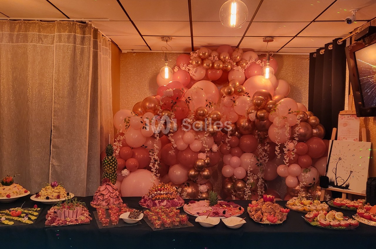 Table garnie de plats variés devant une décoration murale composée de ballons roses et dorés sous un éclairage tamisé.