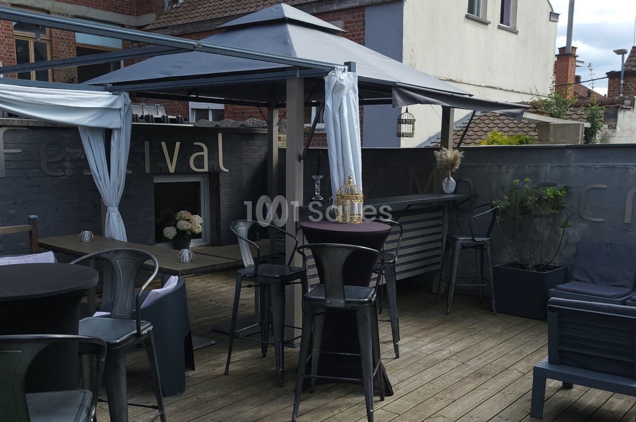 Terrasse en bois avec tables hautes, chaises en métal, pergola et décorations, entourée de murs gris.