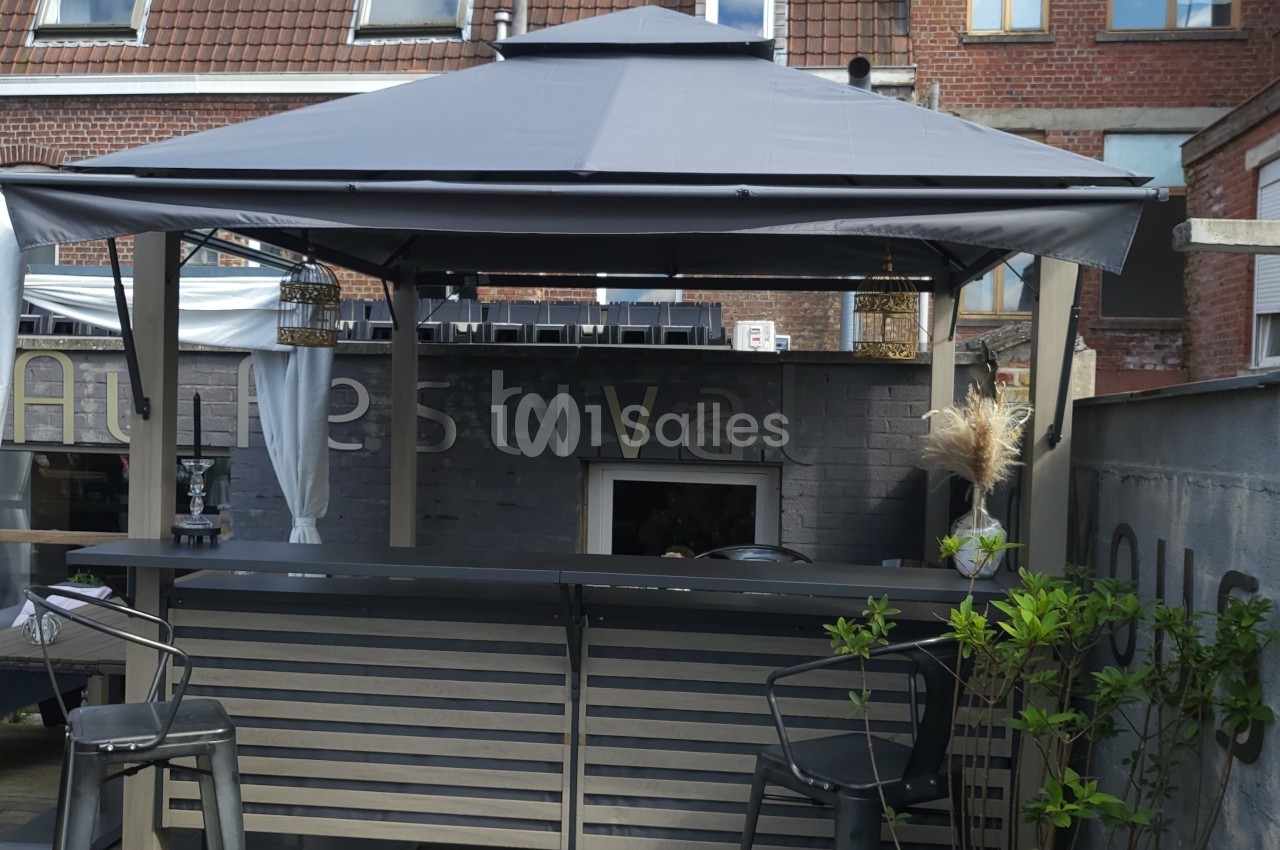 Bar extérieur avec un toit en toile grise, tabourets hauts et décorations végétales, devant un mur en briques.