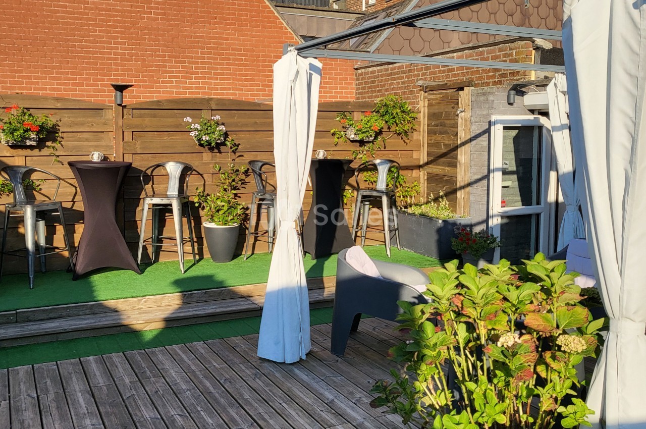 Terrasse ensoleillée avec tables hautes, chaises, plantes en pot et sol en bois et gazon artificiel.