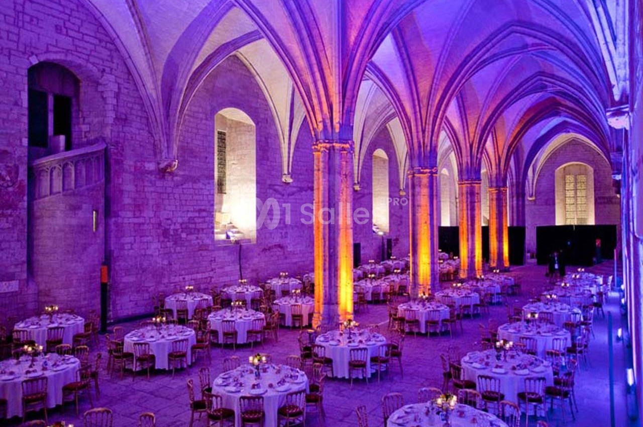 Salle voûtée illuminée en violet et orange, aménagée avec des tables rondes dressées pour un événement.
