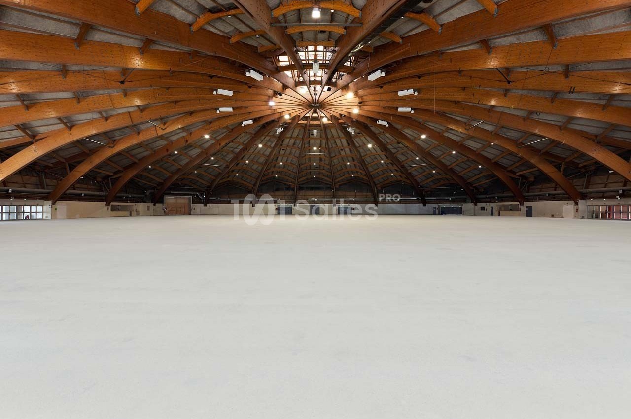 Vue intérieure d'une grande salle avec une structure en bois voûtée et un sol clair, éclairée par des fenêtres en hauteur.