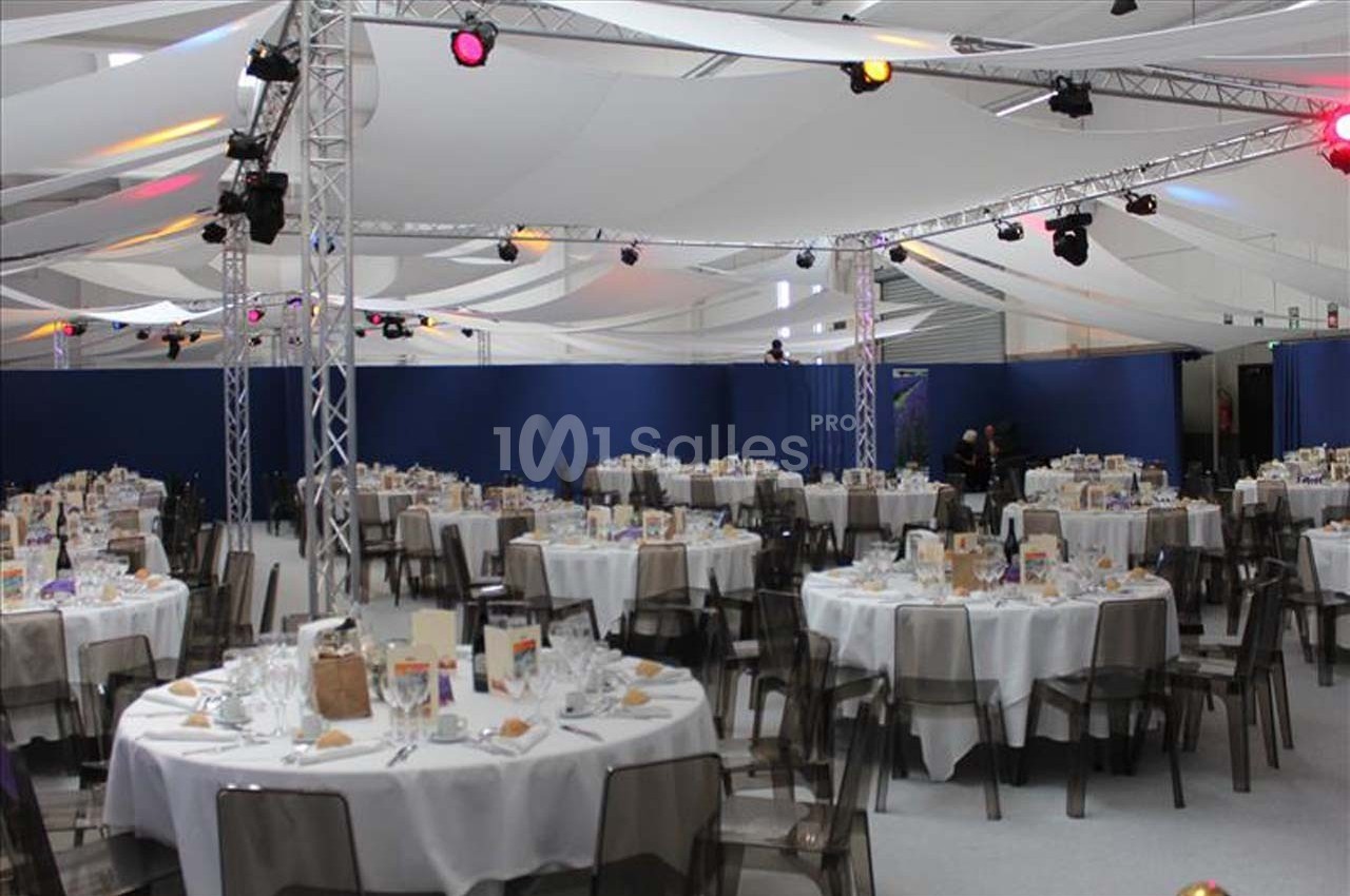 Salle de réception avec des tables rondes dressées pour un événement, sous une structure avec éclairage coloré.