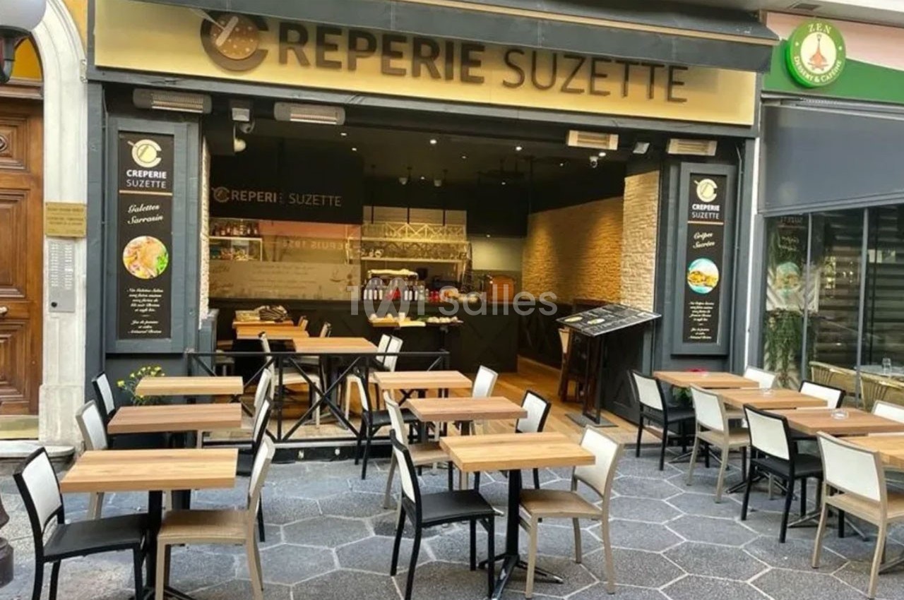 Terrasse d'une crêperie avec tables et chaises en bois, devanture vitrée et enseigne ’Crêperie Suzette’.