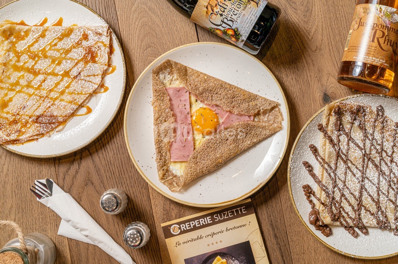 Assiette de galette au sarrasin garnie de jambon, œuf et fromage, entourée de crêpes sucrées et boissons sur une table en…