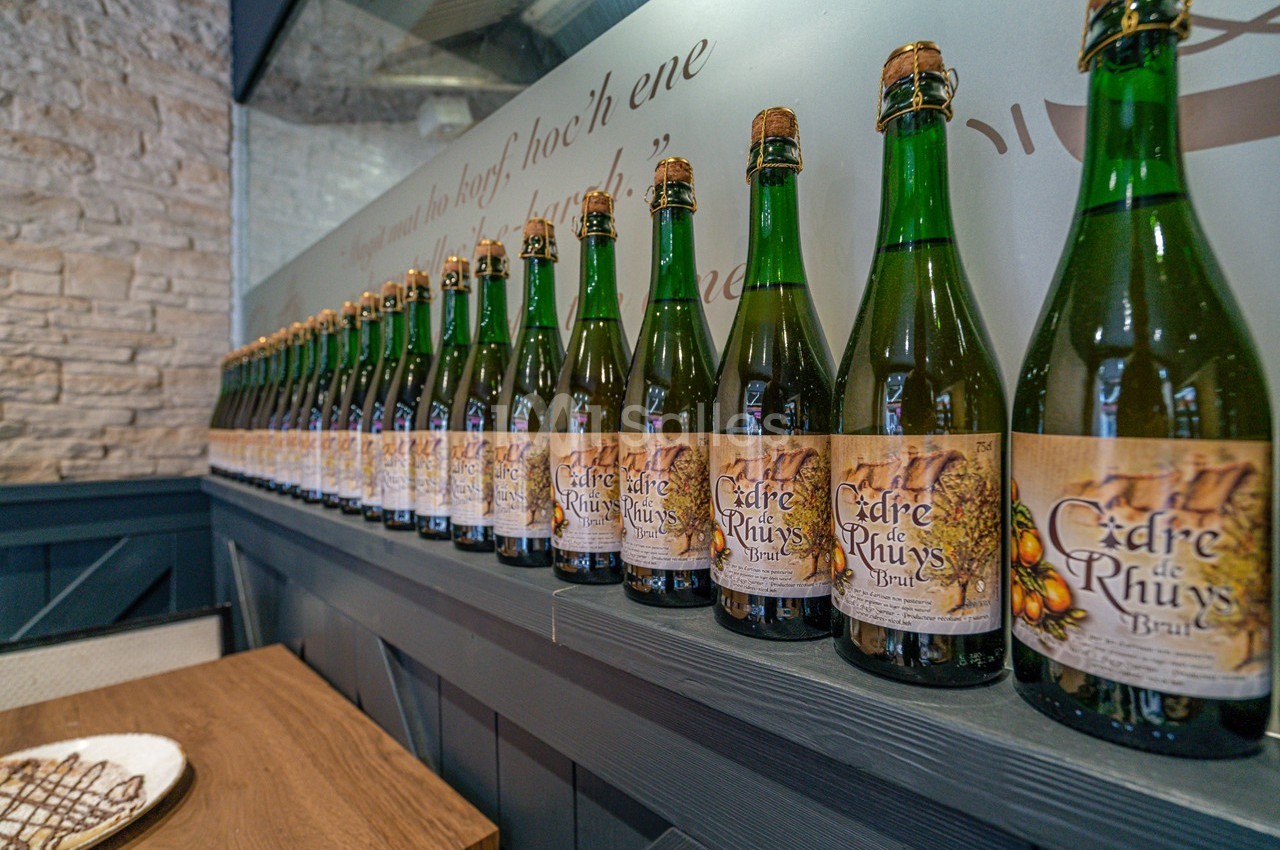 Alignement de bouteilles de cidre sur une étagère en bois, devant un mur en pierre et une vitre décorative.