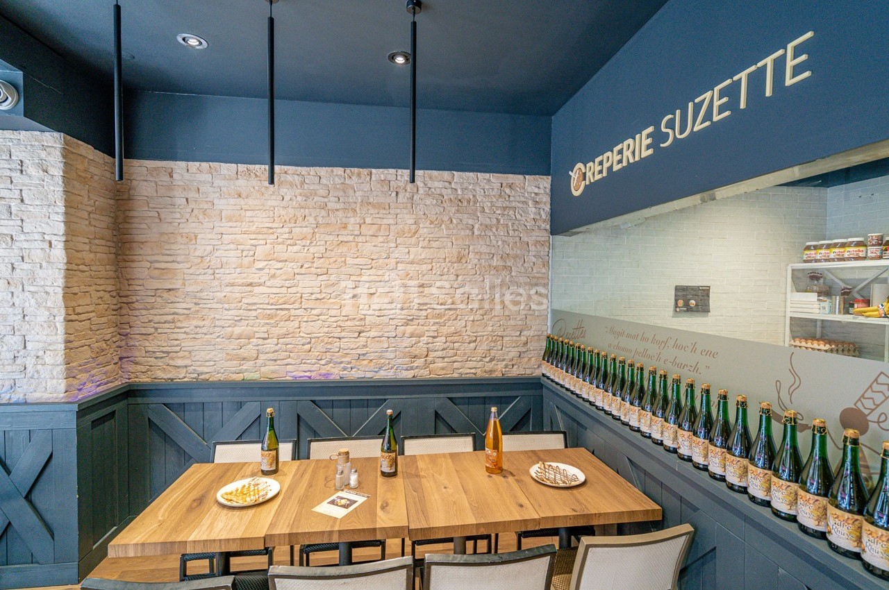 Salle de restaurant avec table en bois, chaises, bouteilles alignées sur une étagère et mur en pierre claire.