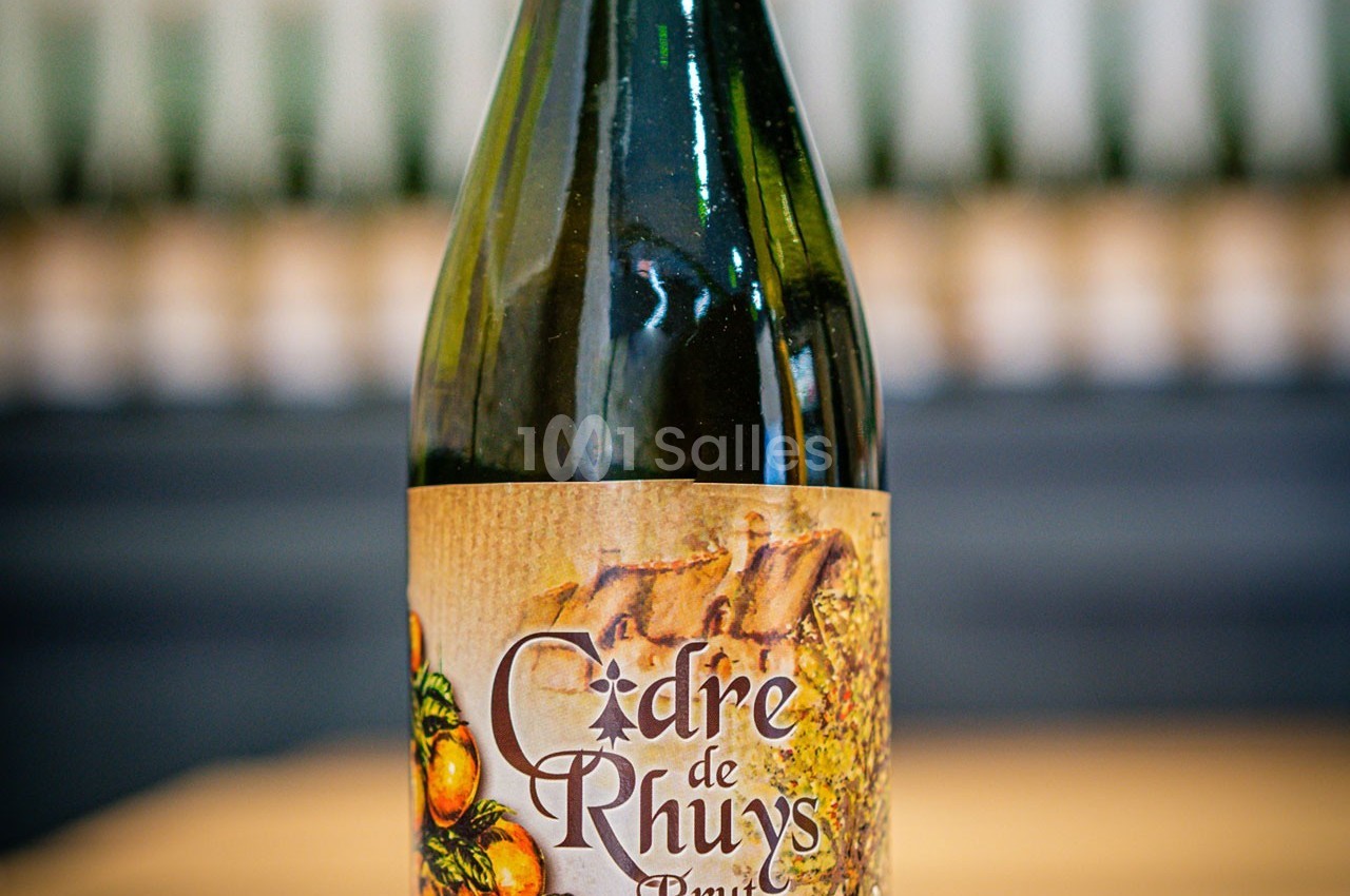 Bouteille de cidre brut avec une étiquette illustrée de pommes et un fond flou de bouteilles alignées.