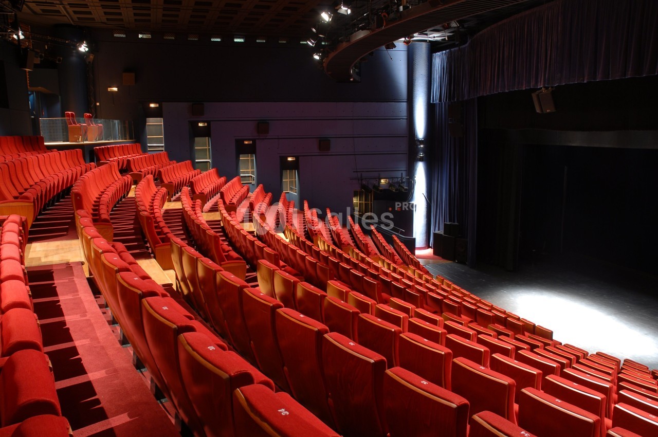 Salle de théâtre vide avec des rangées de sièges rouges et une scène éclairée à droite.