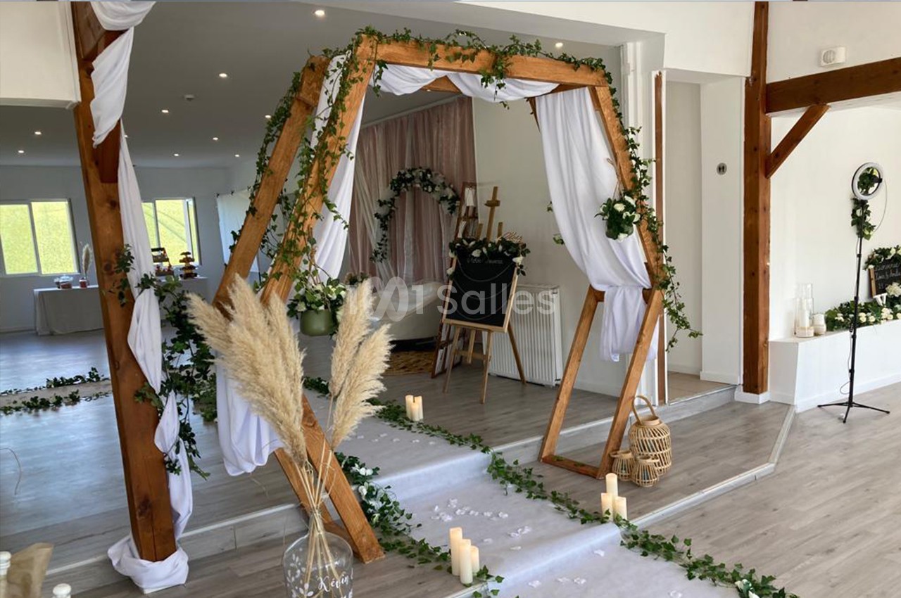 Décoration de cérémonie intérieure avec arche en bois ornée de tissus blancs et feuillages, bougies et pampas au sol.