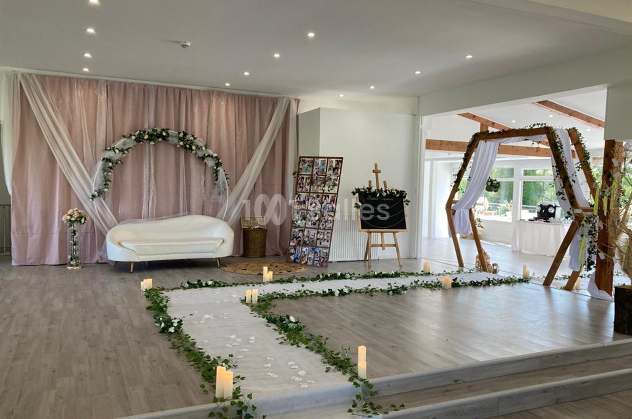 Salle décorée pour un événement avec arche florale, bougies, tapis blanc et panneaux de photos.