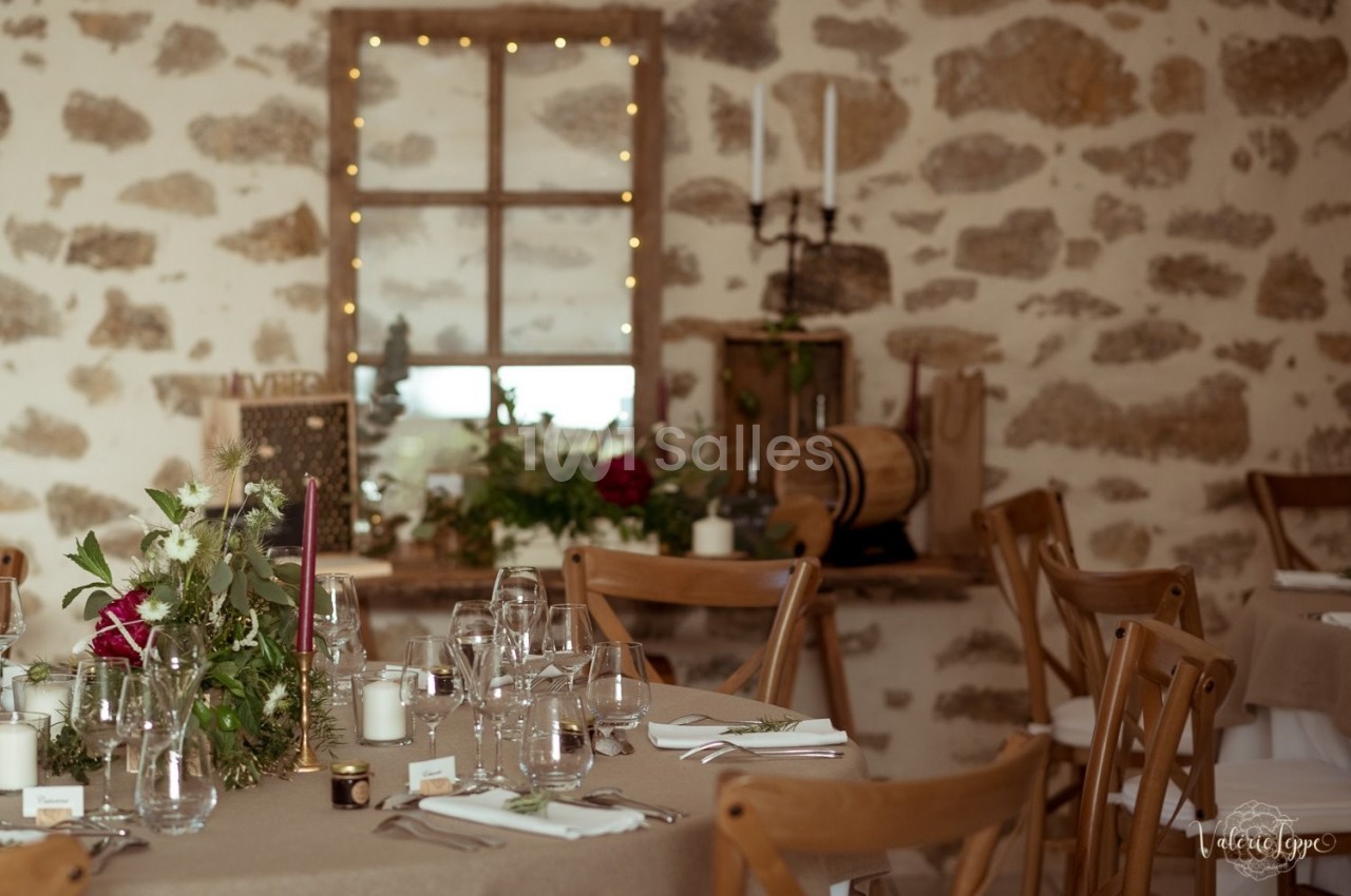 Salle de réception rustique avec tables décorées de fleurs, bougies et vaisselle, devant un mur en pierre.
