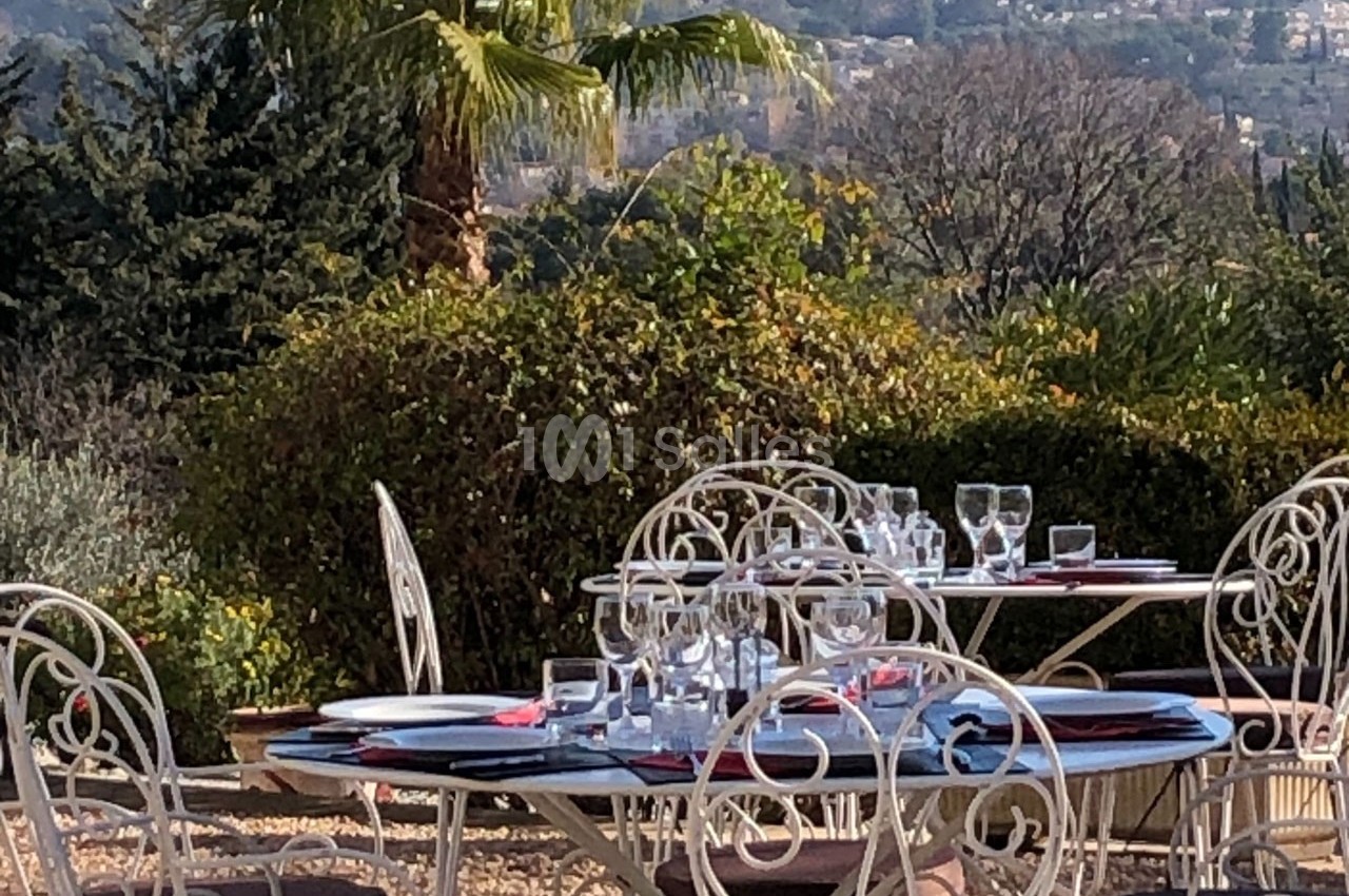 Tables en fer forgé dressées avec vaisselle et verres, disposées en extérieur avec vue sur un paysage verdoyant.
