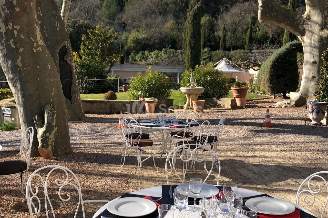 Terrasse extérieure avec tables dressées, chaises en fer forgé, arbres et végétation en arrière-plan.