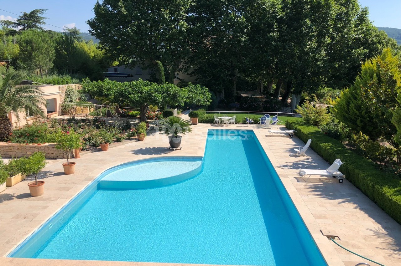 Piscine extérieure entourée de végétation, transats et pots de fleurs sur une terrasse en pierre.