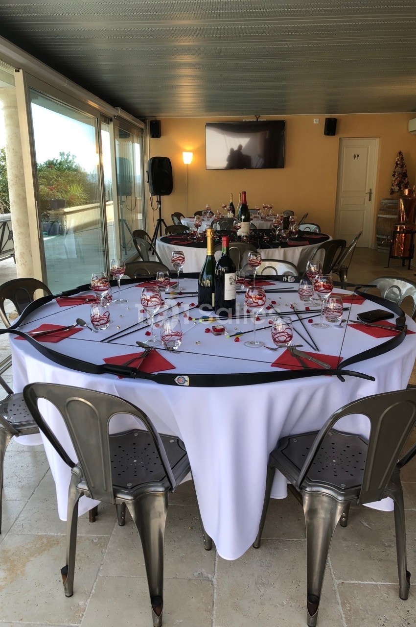 Table ronde dressée avec nappes blanches, verres, serviettes rouges et bouteilles, dans une salle lumineuse avec baies…