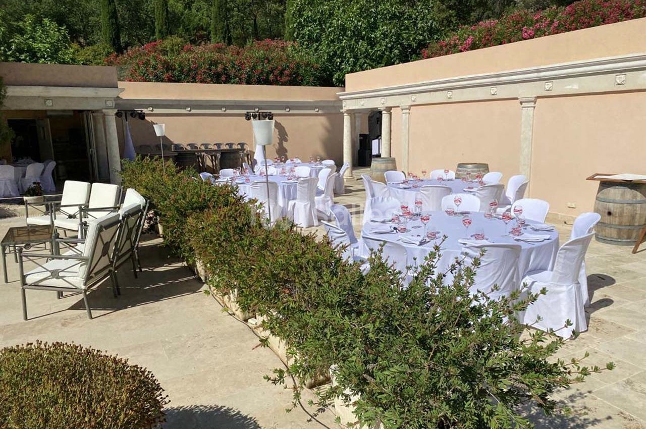 Terrasse extérieure avec tables rondes dressées et chaises blanches, entourée de verdure et de murs en pierre beige.