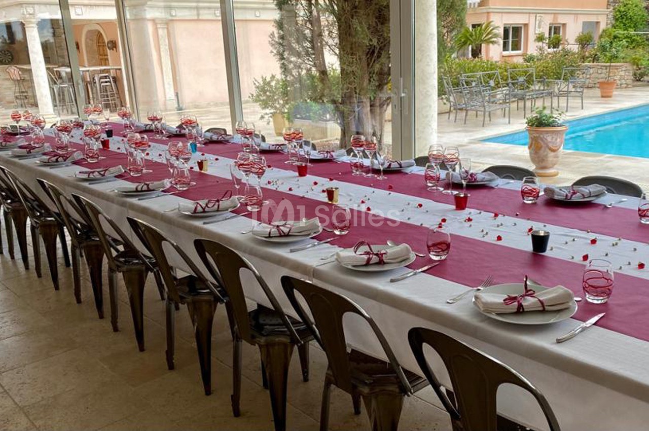 Table longue dressée pour un repas, nappes blanches et bordeaux, vaisselle élégante, près d'une piscine extérieure.