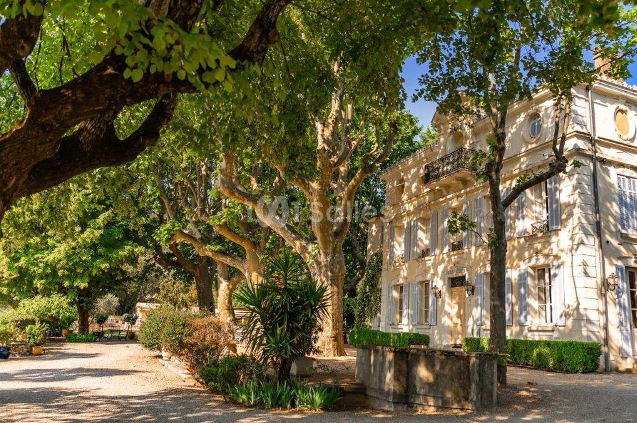 Façade d'une maison blanche entourée d'arbres et de végétation dans un jardin ensoleillé.