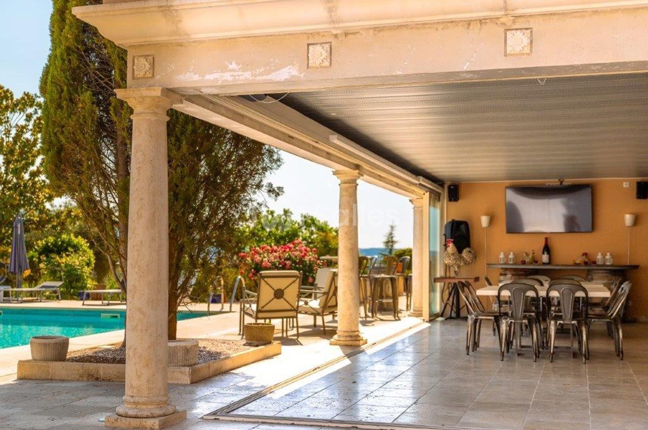 Terrasse couverte avec table et chaises, donnant sur une piscine entourée de végétation et mobilier de jardin.
