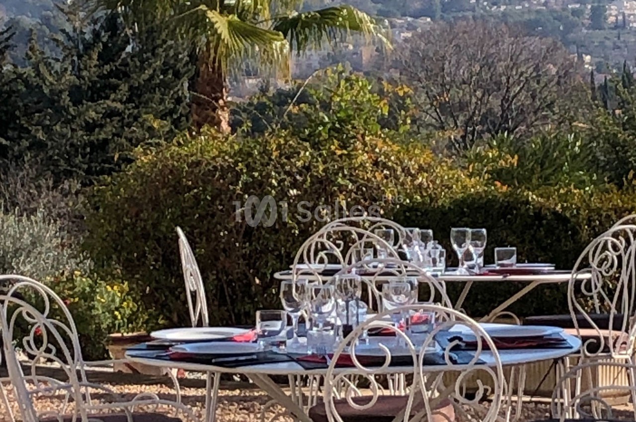 Tables en fer forgé dressées avec vaisselle et verres, situées en extérieur dans un jardin avec vue sur des collines.