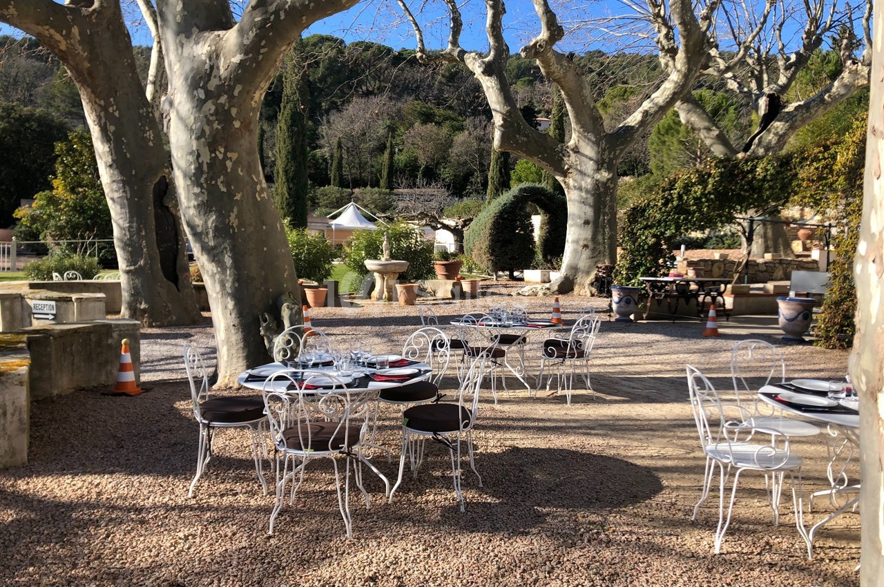 Tables et chaises en fer forgé disposées sur une terrasse en gravier, entourées d'arbres et de végétation.