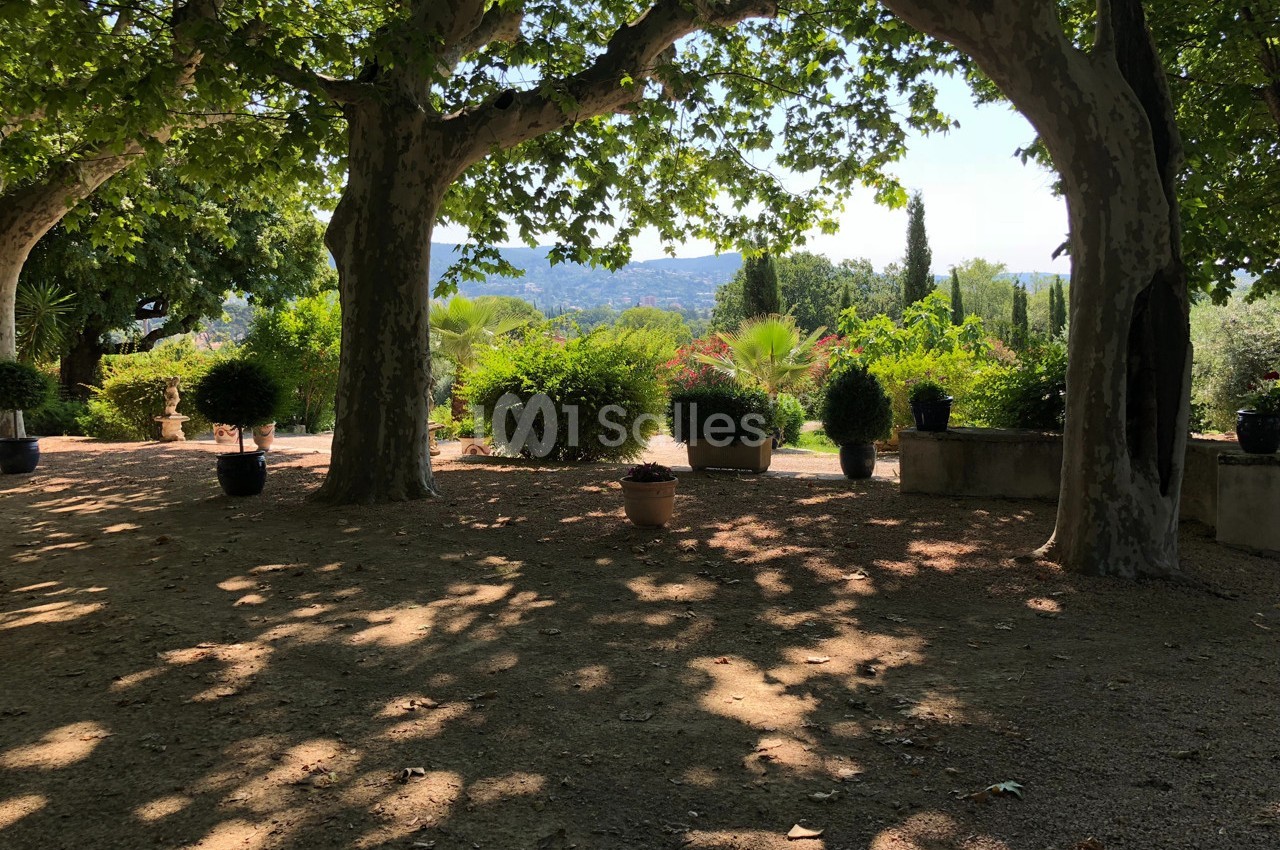 Allée ombragée bordée d'arbres et de plantes en pots, avec vue sur un paysage vallonné en arrière-plan.
