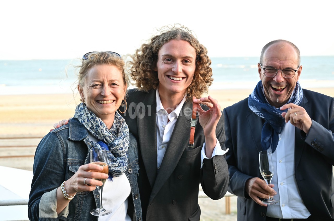 Trois personnes souriantes tenant des verres, posant devant une plage en arrière-plan.